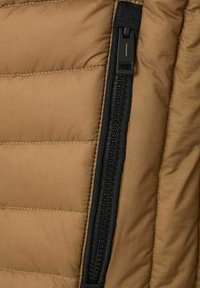 Beige Pufferjacke mit gesteppten Abschnitten. Sie hat einen auffälligen schwarzen Reißverschluss an der Seite, eine strukturierte Oberfläche und kontrastierende Nähte.