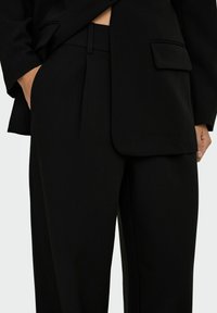 Pantalon noir cintré avec une texture lisse, présentant une coupe droite, des plis et une poche latérale. La ceinture est plate et discrète.