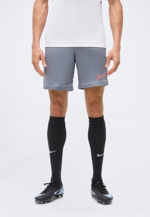 Άνδρας που φοράει γκρι σορτς Nike, μαύρες κάλτσες Nike μέχρι το γόνατο και μαύρα ποδοσφαιρικά παπούτσια, στέκεται μπροστά σε λευκό φόντο.