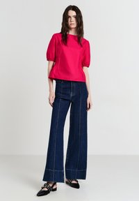 Blusa di raso rosa con maniche a sbuffo corte, abbinata a jeans a gamba larga blu scuro con cuciture visibili e scarpe basse nere con accenti in metallo.