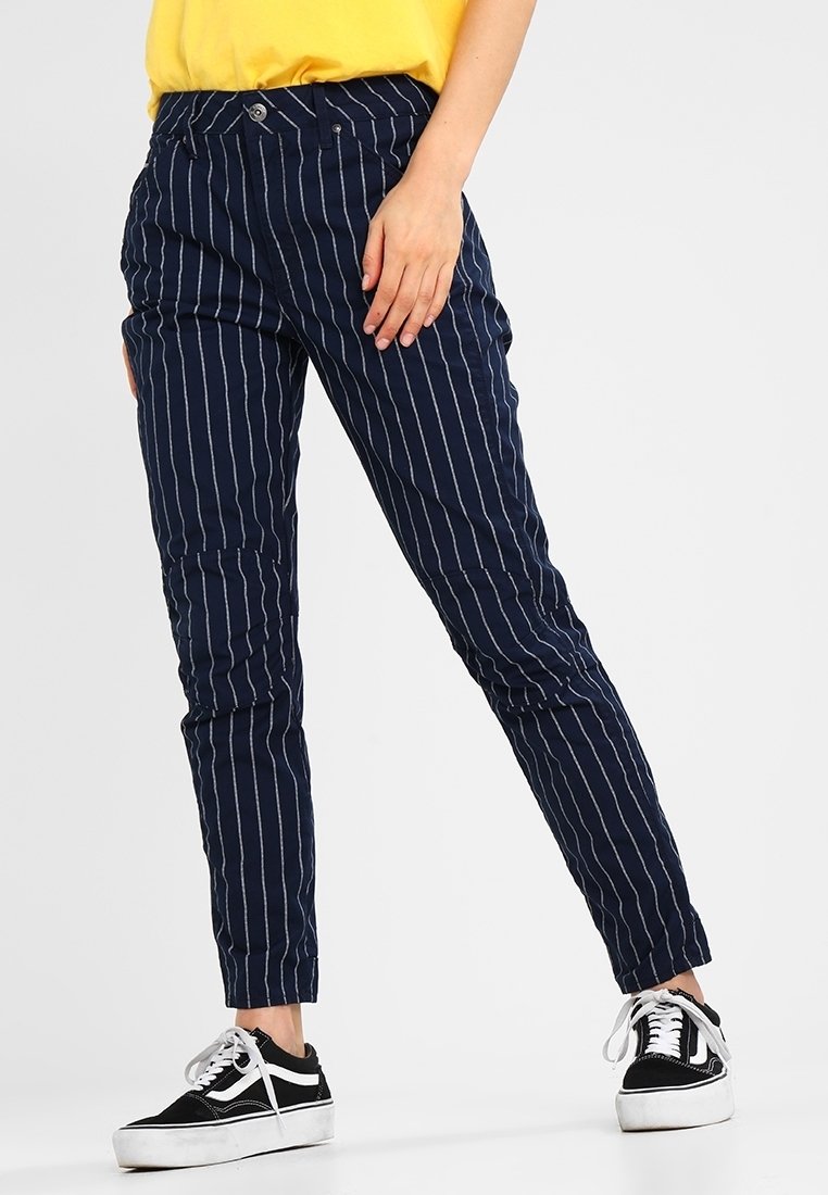 Pantalons navy avec des rayures verticales blanches, présentant un design ajusté, deux poches avant et des ourlets bordés, associés à des baskets noir et blanc.