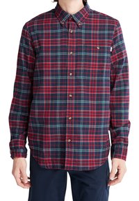 Uomo che indossa una camicia a maniche lunghe con bottoni a quadri rossi, blu navy e verdi, abbinata a pantaloni blu navy scuri, con le mani rilassate ai lati.