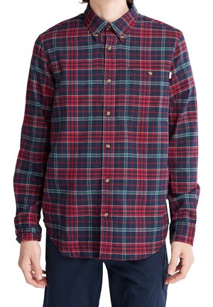 Man in een rood, marineblauw en groen geruit overhemd met lange mouwen, gecombineerd met donker marineblauwe broek, met ontspannen handen naast zich.