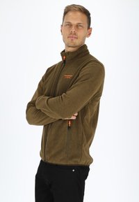 Giacca in pile marrone con zip e colletto alto, dotata di tirazip arancioni e logo ricamato. Indossata sopra pantaloni neri.