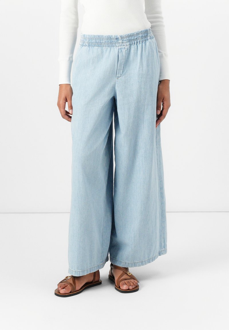 Pantalon large bleu clair avec une taille smockée, fabriqué en tissu en denim. L'ourlet est fini et associé à des sandales plates marron.