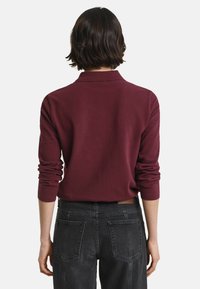 Person with short dark hair wears a long-sleeve burgundy top and high-waisted black jeans, shown from the back against a white background.