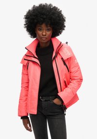 Lys pink polstret jakke med høj krave, lynlåslommer og sorte detaljer. Bæres over en sort sweater og mørke jeans. Glat tekstur.