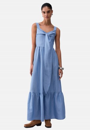 Donna che indossa un abito lungo azzurro senza maniche con dettaglio annodato sul davanti, abbinato a stivaletti marroni e braccialetti d'argento.