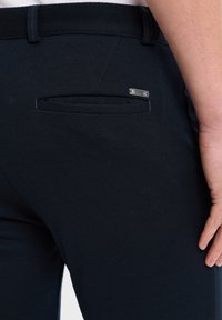 Dunkelblaue Hosen aus strukturiertem Stoff, mit einer einzelnen Gesäßtasche und einem kleinen metallischen Markenetikett am Taschenrand.