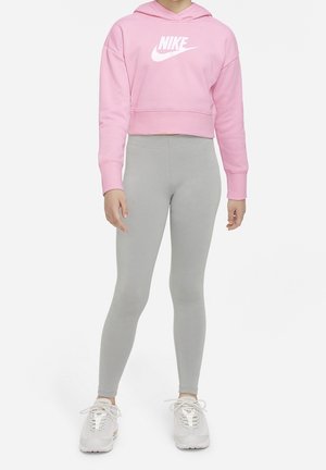 Osoba ubrana w różową krótką bluzę Nike z kapturem, jasnoszare legginsy i białe trampki, stojąca na jednolitym białym tle.