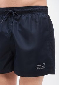 Μπλε σκούρο μαγιό βερμούδα κατασκευασμένο από ελαφρύ υλικό, με ελαστική μέση και κορδόνι και τυπωμένο λογότυπο στο μηρό.