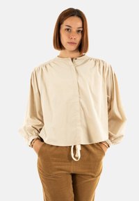 Blusa beige de manga larga con cuello redondo, hombros fruncidos y puños elásticos. La blusa tiene un corte relajado y un dobladillo con cordón.