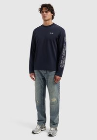 Navy longsleeve shirt met de tekst "Pure Faith" op de linkermouw; gecombineerd met versleten lichtblauwe jeans en metalen sneakers.