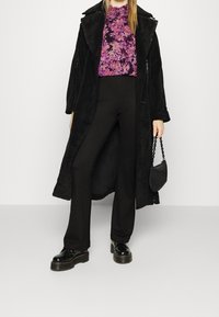 Cappotto in peluche nero su una maglietta floreale viola, abbinato a pantaloni neri e scarpe chunky nere. Piccola borsa nera con tracolla a catena.