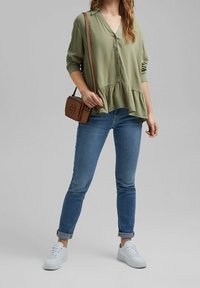 Blusa verde chiaro con ragazzo abbottonato davanti e orlo peplum, abbinata a jeans blu e sneakers bianche. Borsa a tracolla marrone visibile.