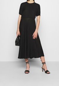 Vestido negro midi con corsé ajustado y falda acampanada, hecho de tela texturizada. Combinado con tacones altos negros con tiras y un bolso.
