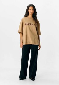 Camelkleurig oversized t-shirt met de graphic "SIMPLICITY" in bordeaux, gedragen met zwarte wijde broek. Gladde stof, ontspannen pasvorm, korte mouwen.