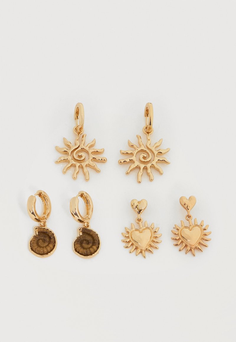 Trois paires de boucles d'oreilles pendantes couleur or avec des spirales solaires, une coquille fossile et un cœur avec des rayons de soleil sur fond blanc.