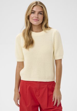 Junge Frau mit langen blonden Haaren, die einen cremefarbenen Strickpullover mit kurzen Ärmeln und hoch taillierte rote Hosen vor einem einfarbigen Hintergrund trägt.