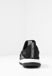 Zapatilla negra de Hugo Boss con suela blanca acolchada, etiqueta en el talón con la marca y etiqueta de tamaño visible dentro del collar, vista desde atrás.
