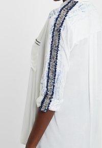 Vit skjorta med blå och silver broderade detaljer längs ärmarna, med textmönster och en avslappnad passform. Texturerad detaljering vid sömmarna.