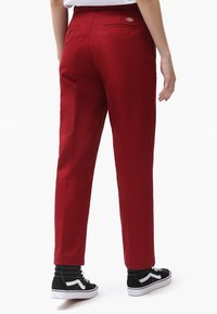 Červené bavlněné kalhoty se rovnýcm střihem, které mají zadní kapsy a visačku s logem. Nosí se s černými vysokými teniskami a vzorovanými ponožkami.