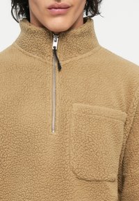 Tan fleece pullover met een hoge kraag, halve rits en een voorkantzak. Geweven stof met een zachte, pluizige uitstraling.