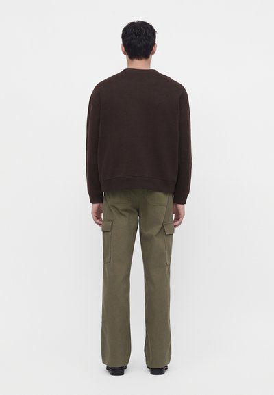 Homme debout de dos, portant un sweat-shirt marron foncé et un pantalon cargo vert olive avec des chaussures noires sur un fond uni.