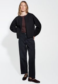 Giacca nera corta con maniche larghe, abbinata a un top marrone testurizzato, jeans neri a gamba larga e scarpe basse marroni scure.