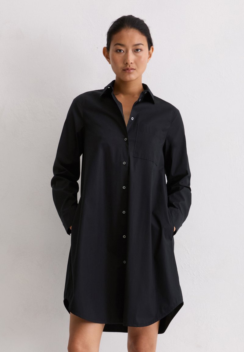 Kobieta stojąca przy białej ścianie, ubrana w czarną sukienkę koszulową z długim rękawem, zapinaną na guziki, z kieszeniami i kołnierzykiem, z rękami w kieszeniach.