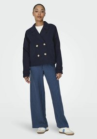 Giacca blu navy doppiopetto con bottoni dorati, caratterizzata da una vestibilità rilassata. Abbinata a pantaloni a gamba larga blu e bianchi a strisce e sneakers bianche.