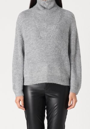 Persoon die een grijze geribbelde pullover draagt met een opstaande kraag met rits en zwarte leren broek, staand tegen een effen achtergrond.