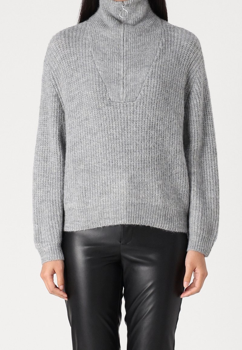 Personne portant un pull gris côtelé avec un col montant zippé et un pantalon en cuir noir, debout devant un fond uni.