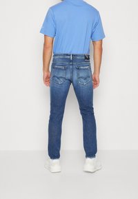 Hombre vestido con jeans azul y zapatillas blancas, de pie con la espalda hacia la cámara frente a un fondo claro y liso.