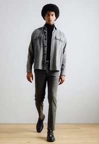 Chemise en flanelle gris à boutons, col roulé noir, pantalons vert olive et chaussures noires ; le modèle se tient devant un fond neutre.