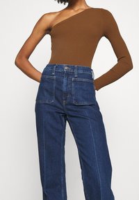 Hnědý žebrovaný body s jedním ramenem a dlouhým rukávem, spojený s vysokými modrými džíny z denimu, které mají přední kapsy a světle šité detaily.