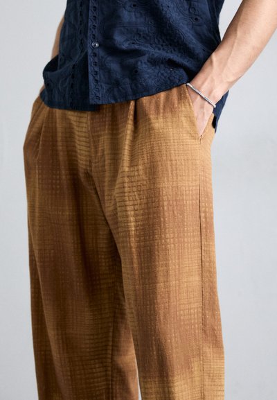 Brown patterned trousers with textured fabric, featuring a relaxed fit and side pockets, partially covered by a dark blue button-up shirt.