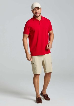 Polo rouge, shorts cargo beiges avec poches latérales et chaussures bateaux marron. La tenue présente une coupe décontractée et une casquette de couleur claire.