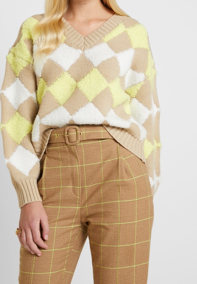 Femme portant un pull à losanges beige, blanc et jaune, associé à un pantalon taille haute marron avec un motif quadrillé jaune et une ceinture assortie.