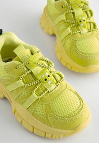 Jaskrawozielone sneakersy mają siateczkową i zamszową teksturę, unikalny system sznurowania oraz wyściełany kołnierz. Gumowa podeszwa z wzorem zapewniającym przyczepność.
