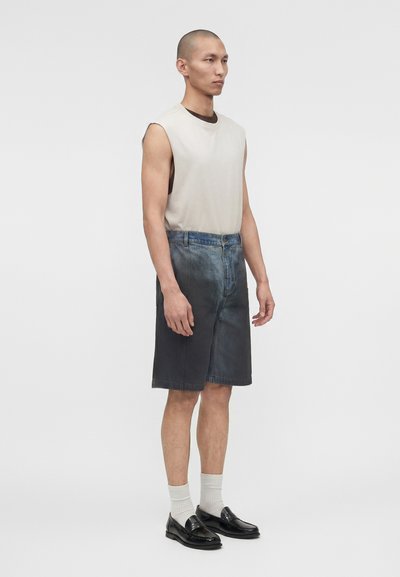 Άνδρας μοντέλο που φοράει ένα μπεζ αμάνικο τοπ, σκούρα τζιν σορτς με διαβάθμιση, λευκές κάλτσες και μαύρα δερμάτινα μοκασίνια, στέκεται μπροστά σε απλό φόντο.