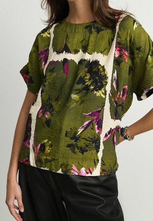 Blouse florale verte à manches courtes, ornée de motifs roses et noirs et d'un tissu texturé. Associée à un pantalon en cuir noir.
