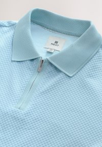 La polo shirt blu chiaro presenta un materiale testurizzato, un collo con zip e un colletto in rete, con un'etichetta che mostra il marchio "NEXT" e la taglia "M."