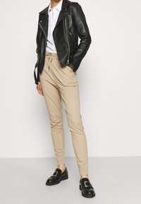 Person som bär beige dragsko-byxor, svart läderjacka, vit skjorta och svarta loafers med silverknäppning, står mot en enfärgad bakgrund.