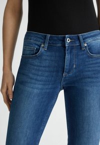 Pantalones vaqueros de mezclilla azul con un diseño ajustado, estilo de cinco bolsillos y un ligero efecto de desgarro. La cintura cuenta con un cierre de botón metálico.