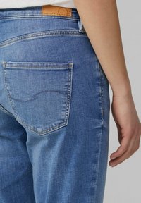 Jeans en denim bleu clair avec une poche arrière, des coutures visibles et une étiquette en cuir marron au niveau de la taille. Texture lisse, légèrement décolorée.