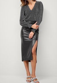 Metallic graues Langarmoberteil mit V-Ausschnitt, kombiniert mit einem hochsitztenden Rock aus schwarzem Kunstleder, der einen Schlitzausschnitt bis zum Oberschenkel hat. Silberschuhsandalen mit Absatz.