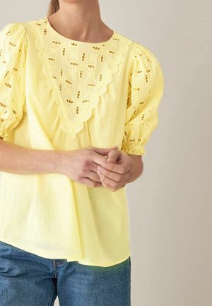 Femme portant un chemisier jaune avec des détails en dentelle ajourée festonnée et des manches bouffantes, associé à un jean bleu en denim.