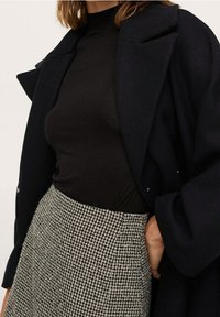 Zwarte lange mouwtop onder een marineblauwe jas, gecombineerd met een rok in houndstoothpatroon. De jas heeft een gestructureerde kraag en knopen.