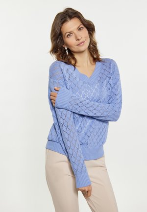 Hellblauer Strickpullover mit Rautenmuster, V-Ausschnitt und gerippten Bündchen und Saum. Kombiniert mit beigen Hosen.